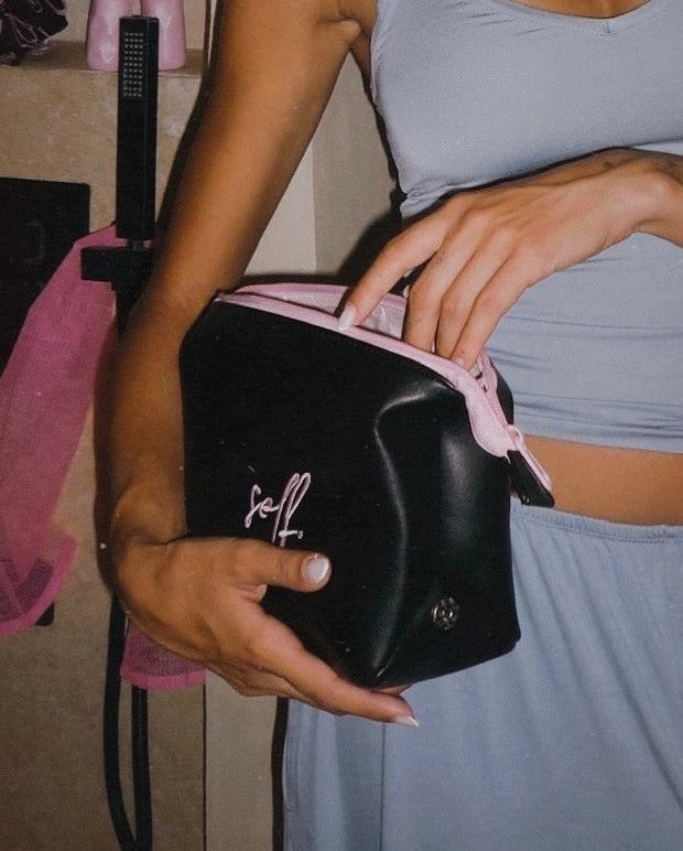 Person holding a black handbag with 'self' branding in a casual setting.