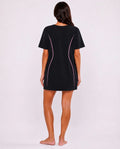 A woman shown from the back wearing a black short-sleeve t-shirt dress with pink curved piping details, standing against a light gray studio background.