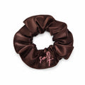 A dark brown satin hair scrunchie with a pink embroidered script logo on the front, displayed on a clean white background.