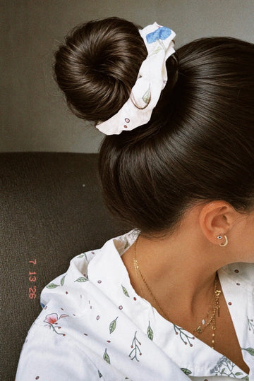 A close-up lifestyle shot of a woman from the back and side, wearing a white fabric hair scrunchie with a blue bow and small printed details, paired with a white patterned shirt.