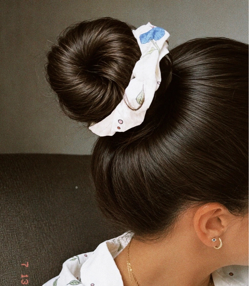 A close-up lifestyle shot of a woman from the back and side, wearing a white fabric hair scrunchie with a blue bow and small printed details, paired with a white patterned shirt.