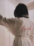 A woman viewed from behind wearing a cream-colored robe with 'Bride' embroidered in pink script on the back, standing in front of a white paneled door.