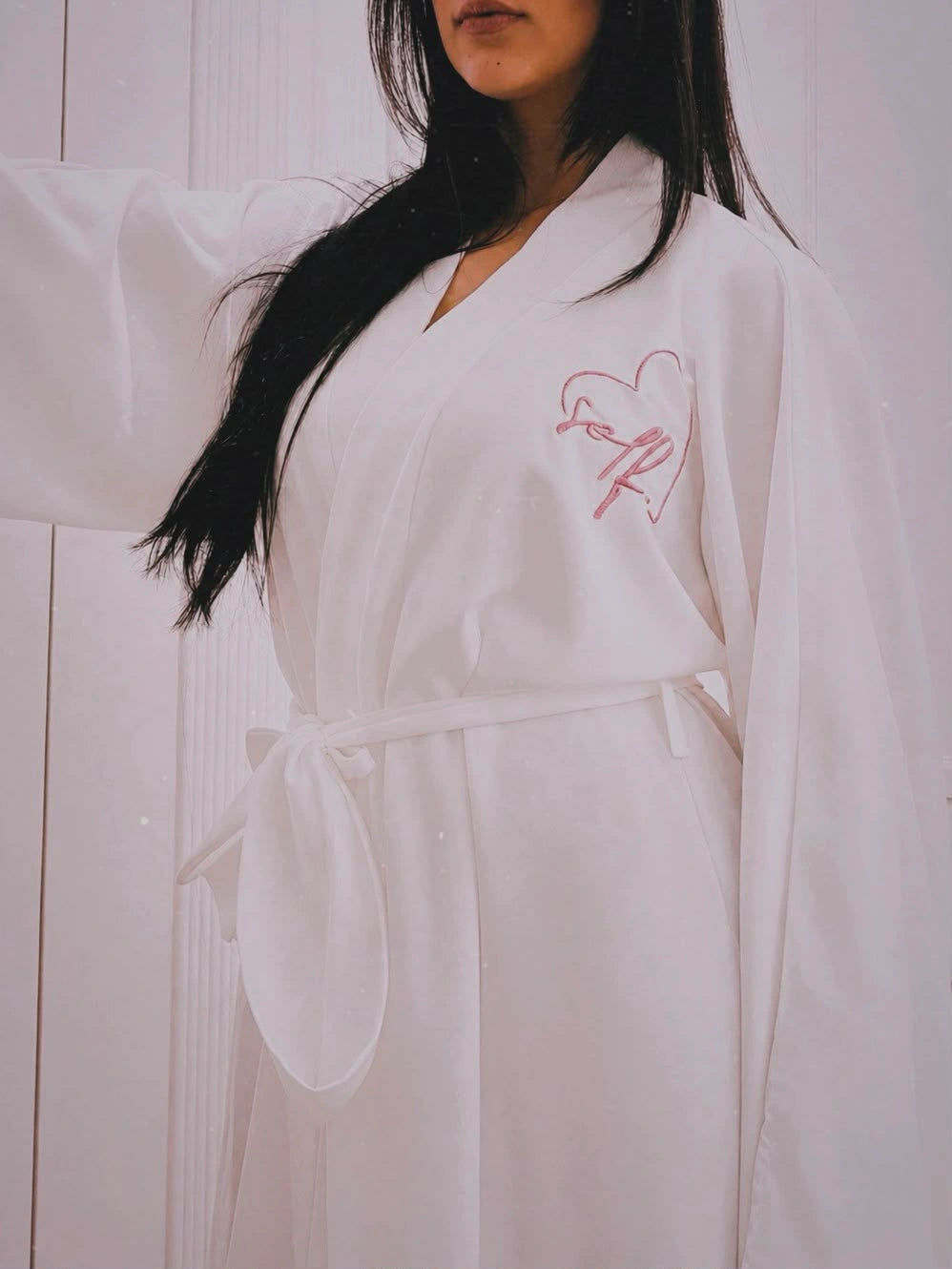 A woman wearing a white robe with a pink embroidered heart and script design on the chest, standing against a white paneled background.
