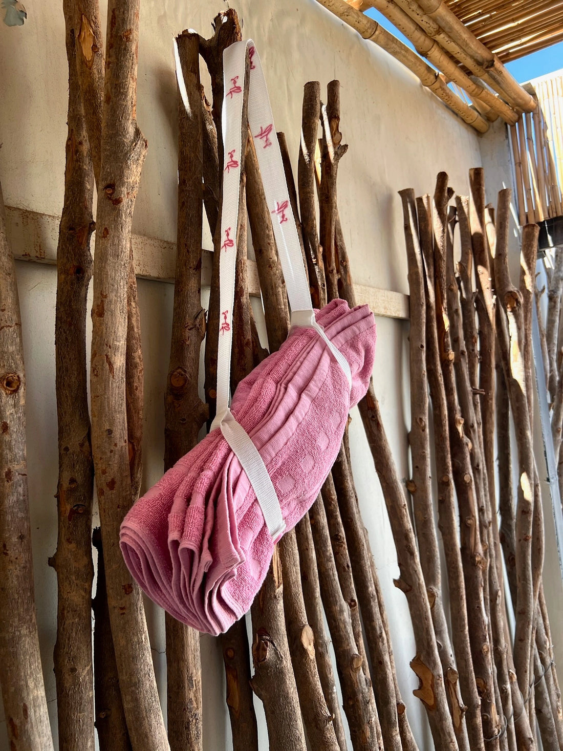 Pink towel wrapped in twine hanging among wooden sticks against a light-colored wall.