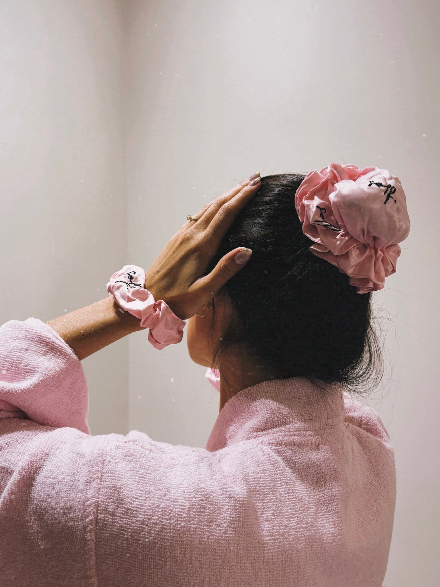A person with dark hair wearing a large pink fabric scrunchie and a matching pink scrunchie on their wrist, seen from behind while wearing a pink textured robe.