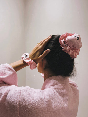 A person with dark hair wearing a large pink fabric scrunchie and a matching pink scrunchie on their wrist, seen from behind while wearing a pink textured robe.