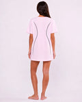 A woman viewed from behind wearing a light pink short-sleeve t-shirt dress with black curved line details on the back, standing against a plain white studio background.