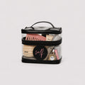 A clear zippered cosmetic bag with a black handle and trim, filled with makeup items including a pink comb, cream products, and a round black lid with pink script branding, photographed on a light gray background.