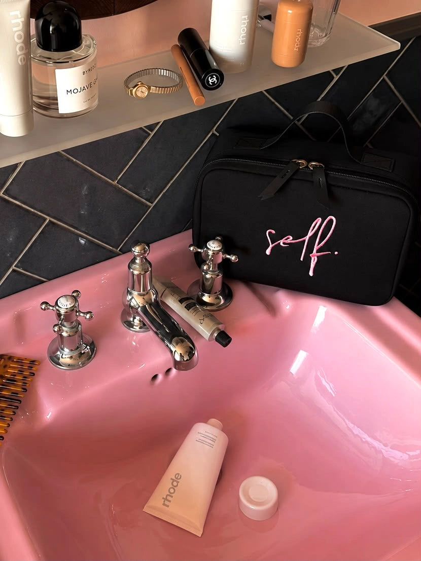 A black zippered cosmetic bag with pink script text reading 'self.' 