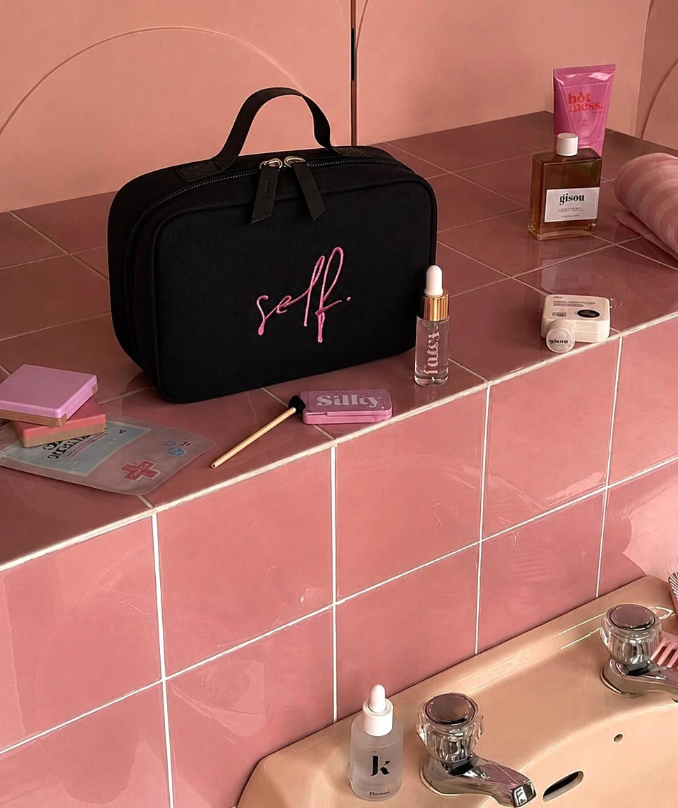 A black cosmetic bag with pink script lettering reading 'self.' 