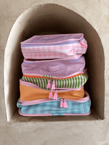 A stack of five colorful zippered pouches or small bags placed inside a rustic beige stone alcove. The bags feature pastel patterns including pink and white gingham, lavender, green and white stripes, solid orange, and light blue and white gingham, each with pink zipper pulls shaped like small figures.