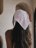 A woman viewed from behind wearing a white bandana with pink paisley patterns tied around her head, set against a rustic beige wall with a wooden archway.