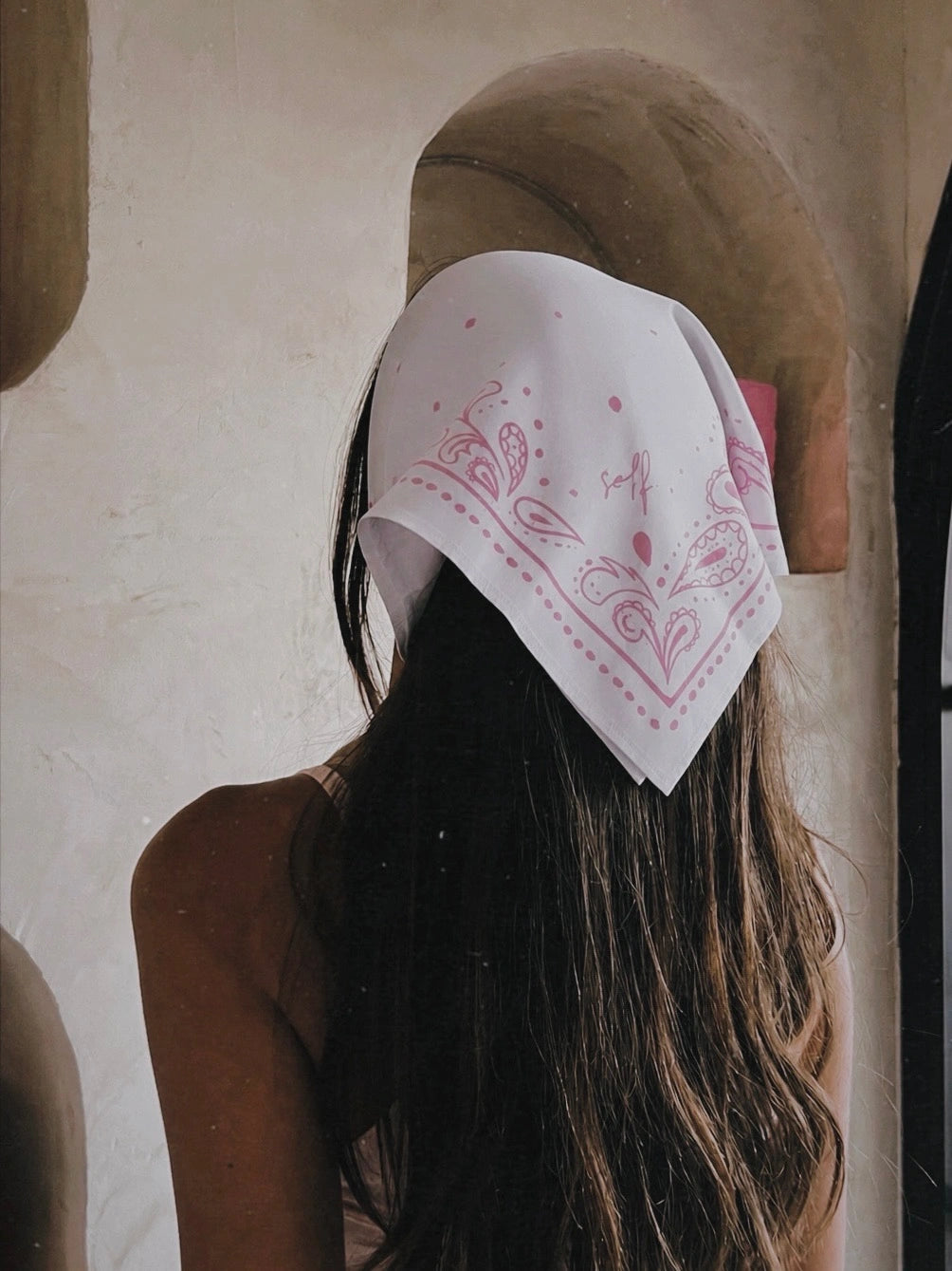 A woman viewed from behind wearing a white bandana with pink paisley patterns tied around her head, set against a rustic beige wall with a wooden archway.