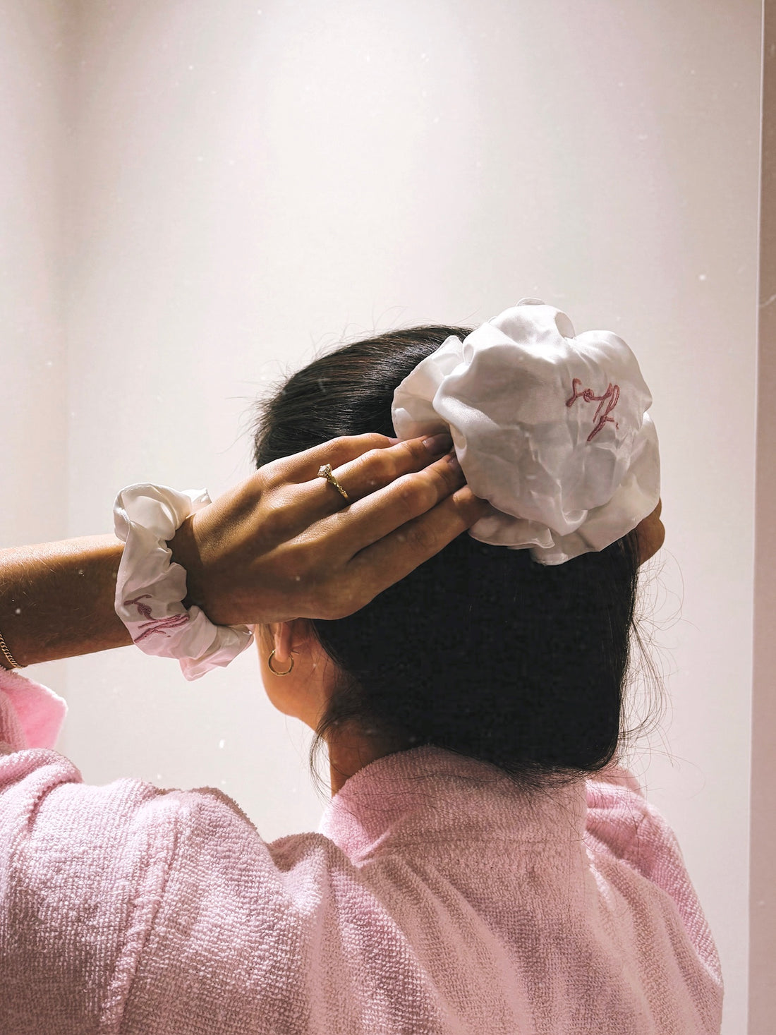A person with dark hair wearing a large pink fabric scrunchie and a matching pink scrunchie on their wrist, seen from behind while wearing a pink textured robe.