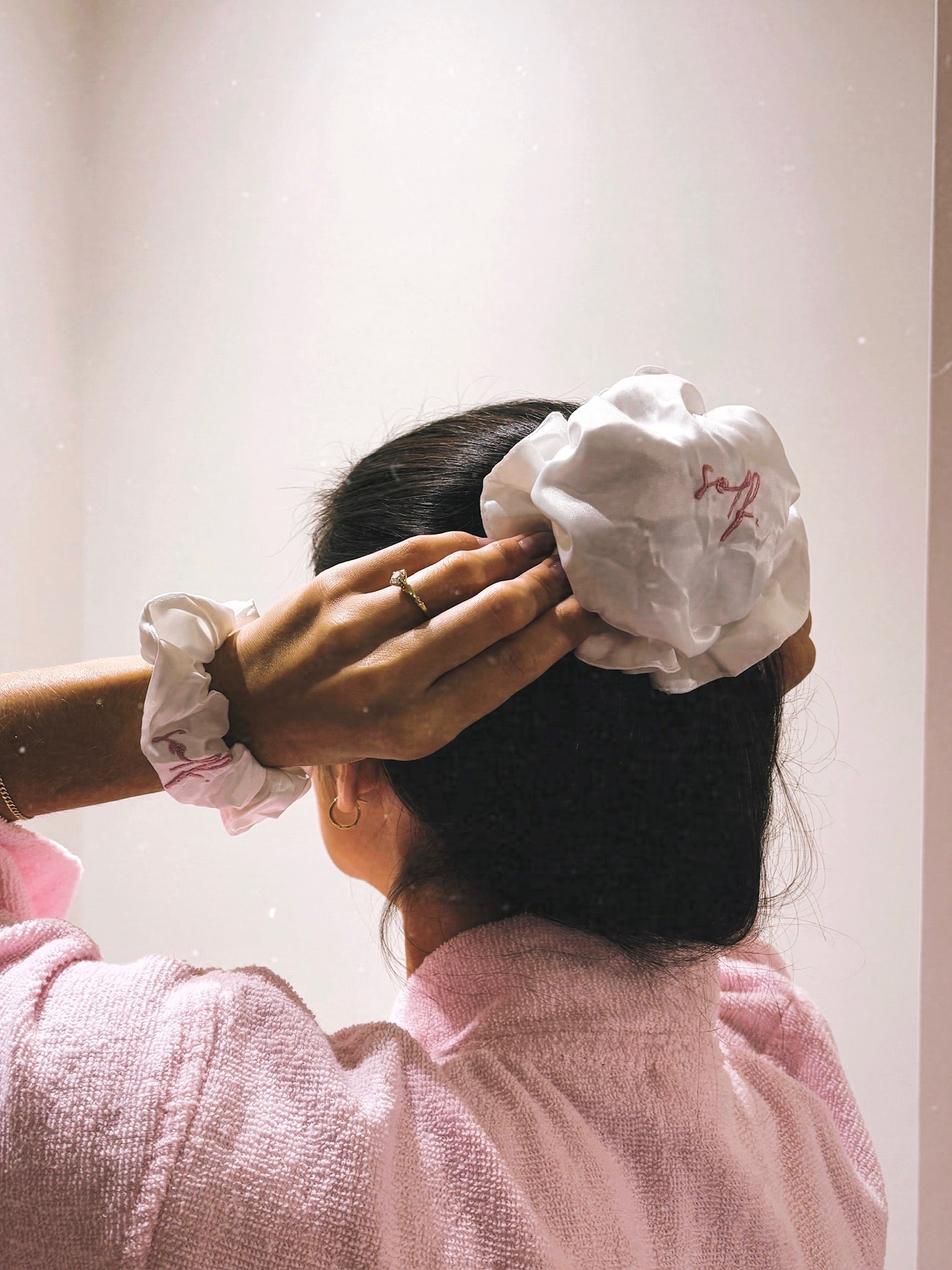 A person seen from behind wearing a large white fabric scrunchie in their dark hair, with a matching light pink scrunchie visible on the other shoulder, against a plain light background.